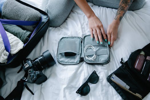 Woman with tattoos packs electronics, camera, and sunglasses in a suitcase on a bed.
