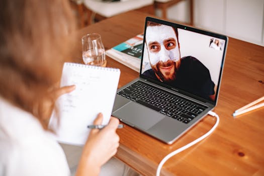 A man in a facial mask on a video call with a woman taking notes by a laptop.